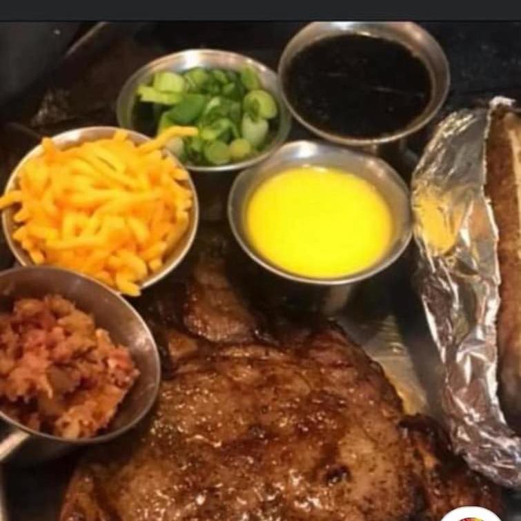 A grilled steak plated with a foil-wrapped baked potato and bowls of toppings — cheese, green onions, and bacon bits