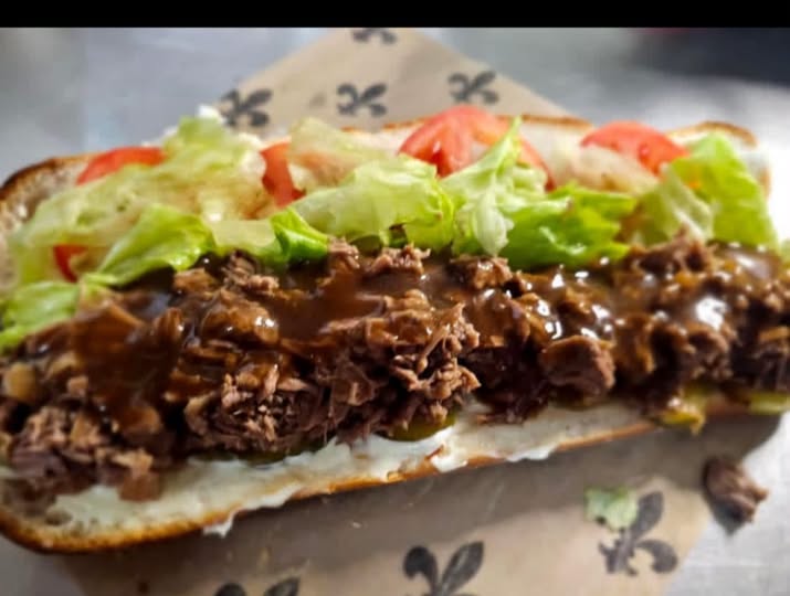 A dressed roast beef poboy piled with gravy-soaked meat, lettuce, and tomato on a long roll